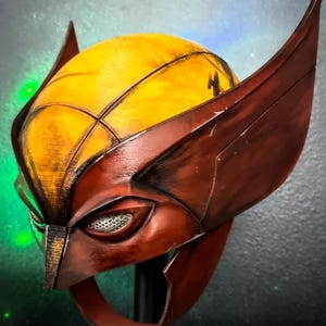 May include: A yellow and dark red helmet featuring large, pointed side fins and a central ridge. The helmet has a textured finish with visible layer lines, almond-shaped eye openings with white mesh inserts, and a chin strap extending downward.