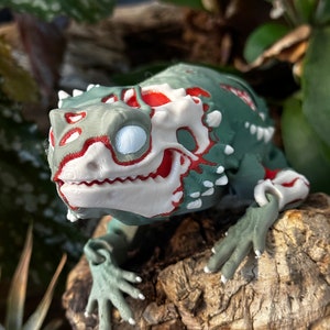 May include: A 3D printed green and red lizard figurine with white details. The lizard is posed on a piece of wood with its head tilted to the side.