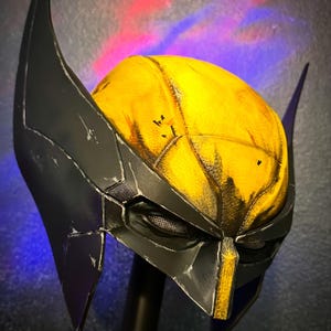 May include: A yellow and black superhero mask featuring tall, pointed side fins, a textured yellow dome with weathered detailing, and a black faceplate with mesh eye coverings and a central vertical yellow stripe, presented on a display stand.