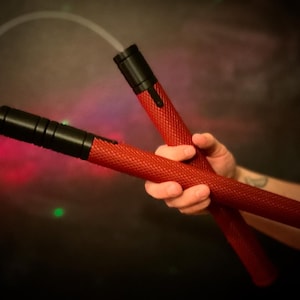 May include: A pair of red and black nunchucks with a textured grip. The nunchucks are held in a person's hand.