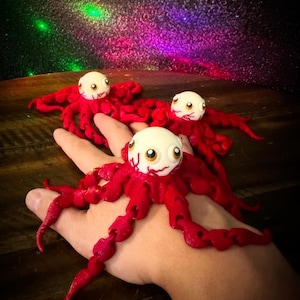 May include: Three red octopus figurines with white and tan eyes and a detailed tentacle design. The octopus figurines are on a wooden surface with a colorful, starry background.
