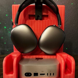 May include: A red 3D printed stand with a slot for headphones and a space for a small computer. The stand has a textured surface and a black headphone stand. The computer has a silver finish and ports on the back. The stand is on a wooden surface.