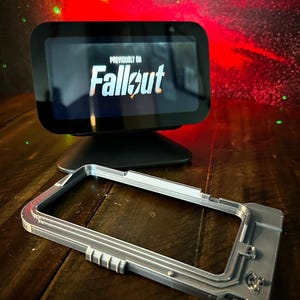 May include: A silver-colored 3D printed case for a smart home device. The case has a textured surface and a raised design. The case is sitting on a wooden table. The screen of the device shows the text "Previously on Fallout".