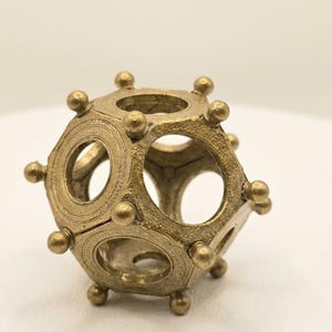 Puede incluir: Un objeto dodecaedro de color oro metálico que presenta una estructura geométrica hueca con aberturas circulares en cada cara. Los bordes están adornados con pequeñas protuberancias esféricas en cada vértice y la superficie tiene un acabado texturizado.