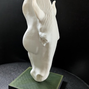 Könnte beinhalten: Weiße 3D-gedruckte Pferdekopfskulptur mit grünem Sockel. Die Skulptur steht auf einer schwarzen Oberfläche.