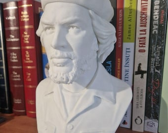 May include: A white bust sculpture of a man wearing a beret and a collared shirt. The sculpture has a detailed face with a beard and is displayed on a wooden shelf in front of a row of books.