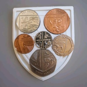 Puede incluir: Un expositor blanco en forma de escudo con seis monedas británicas. Las monedas están dispuestas en círculo con la moneda de cinco peniques en el centro. Las monedas son de diferentes denominaciones, incluyendo dos peniques, cinco peniques, diez peniques y cincuenta peniques. Las monedas son todas de color plateado con un centro de color cobre.