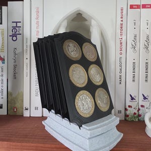 Puede incluir: Un expositor negro para monedas con una base gris, que contiene varias monedas. El expositor está colocado sobre una estantería de madera, con libros al fondo. Las monedas son visibles.