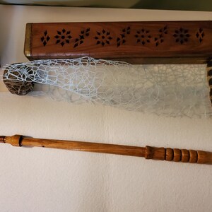 May include: A wooden box with a carved design and a wooden wand with a black tip. The box is closed and has a light brown finish. The wand is about 12 inches long and has a dark brown finish. The wand is lying on a white surface.