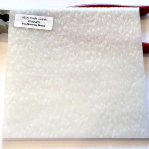 May include: A close-up of a white, textured, square tile with a label that reads "Wispy White Granite Wismach From Mixed Bag Mosaics".