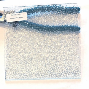 May include: Clear glass sheet with a confetti-like texture. The glass has a bubbly pattern and is accented with two teal-colored lines. A label in the upper left corner reads "Confetti Clear From Mixed Bag Mosaics."