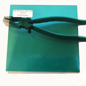 May include: A square sheet of aqua-blue/green stained glass with a pair of green handled pliers resting on top. A white label with black text is affixed to the glass. The glass is likely for mosaic art or stained glass projects.