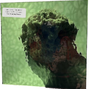 May include: Sage green transparent glass with a textured surface. The glass has a cloudy, layered appearance with a mix of green, brown, and blue hues. A white label in the upper left corner reads "Sage Green Transparent English Muffle Wissmach From Mixed Bag Mosaics."