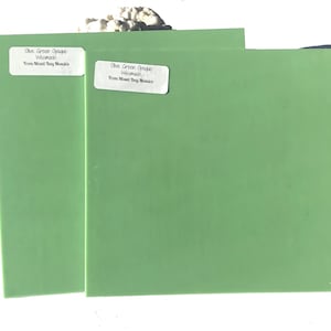 May include: Two olive green, opaque, rectangular tiles. Each tile has a white label with the text "Olive Green Opaque Wissmach From Mixed Bog Mosaics". The tiles are displayed on a white surface with a small pile of white stones.