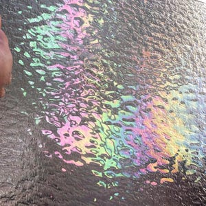 May include: Close-up of a dark, textured surface with iridescent oil slick patterns. The oil creates a rainbow effect with swirls of green, pink, yellow, and blue. The surface appears to be slightly wet, reflecting the light.