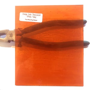 May include: An orange-colored, transparent glass tile with the text "Orange Soda Transparent Tiffany Today From Mixed Bag Mosaics" on a white label. A pair of black-handled pliers is partially visible.