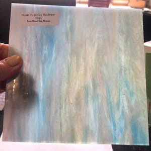 May include: A square piece of stained glass with a wispy pattern in shades of purple, sky blue, and amber. The glass has a translucent quality, with the colors blending together. The text on the label reads: "Medium Purple/Sky Blue/Amber Wispy From Mixed Bag Mosaics."
