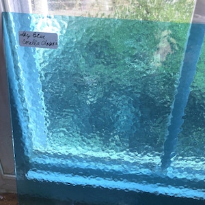 May include: A sheet of textured, sky blue glass. The glass has a rippled surface, creating a water-like effect. A white label in the upper left corner reads "Sky Blue Corella Classic" in handwritten script. The glass is translucent, allowing light to pass through.