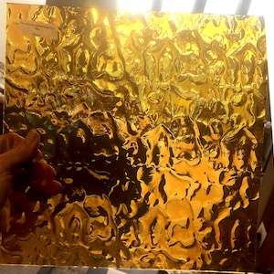 May include: A sheet of textured, golden-colored glass. The surface has a bubbly, irregular pattern, catching the light and creating a shimmering effect. The glass is held by a hand, showcasing its reflective qualities.
