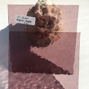 May include: Two rectangular, translucent glass sheets in a lilac hue, with a textured surface. A white label with black text is visible. A light-colored, textured material is behind the glass.