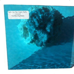 May include: A close-up of a teal blue glass tile with a white label that reads "North Sea Blue English Muffle Wissmach From Mixed Bag Mosaics".