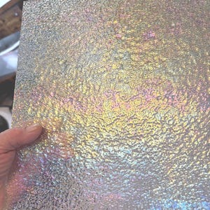 May include: A square piece of iridescent textured glass. The glass has a rough, bubbly surface that reflects a spectrum of colors, including yellow, pink, and blue. The glass is held by a hand, showcasing its reflective qualities.