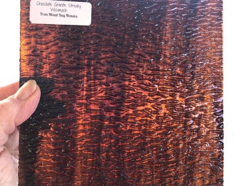 Puede incluir: Una lámina de vidrio estriado marrón y naranja con una superficie texturizada. El vidrio está etiquetado como "Chocolate Granite Streaky Wissmach From Mixed Bag Mosaics".