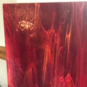 Può includere: Un foglio rettangolare di vetro colorato caratterizzato da un motivo vibrante e vorticoso di tonalità rosso cremisi intenso e arancione brillante. La superficie presenta una consistenza traslucida e marmorizzata con striature organiche.
