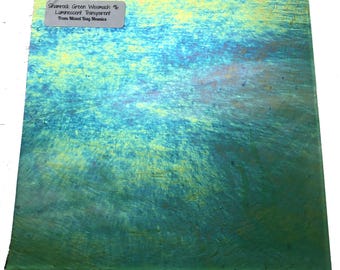 May include: A square piece of iridescent glass with a color gradient of green, blue, and yellow. The glass has a textured surface and a label that reads "Shamrock Green Wismach % Lumnescent Transparent From Mixed Bag Mosaics."