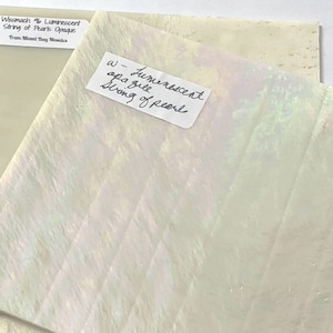 May include: A sheet of iridescent glass with a white background and a subtle green hue. The glass has a string of pearls pattern and is labeled "Wissmach 96 Luminescent String of Pearls Opaque" and "a-Luminescent apa gile String of pearl".