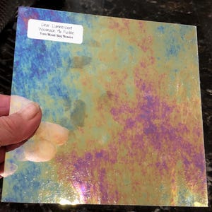 May include: A square piece of clear, iridescent glass held by a hand. The glass has a mottled pattern of blue, yellow, and purple hues. A white label with black text reads "Clear Luminescent Wissmach 96 Fusible From Mixed Bag Mosaics."