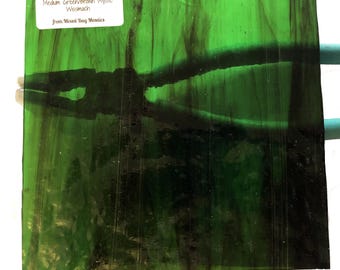May include: A square piece of stained glass in shades of green and brown. The glass has a textured appearance with dark, organic patterns. A white label with black text is affixed to the top left corner, reading "Medium Green/Brown Mystic Wissmach".