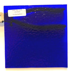 May include: Cobalt blue stained glass square with a textured surface. The glass has dark streaks and a white label with text that reads "Cobalt Blue Corella Wismach" and "From Mount Ring Mosaics".