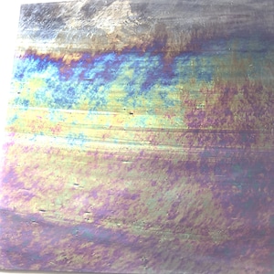 May include: A close-up of a metallic surface with iridescent colors, showing a rainbow effect with shades of blue, green, purple, and gold. The surface has a textured, brushed finish.