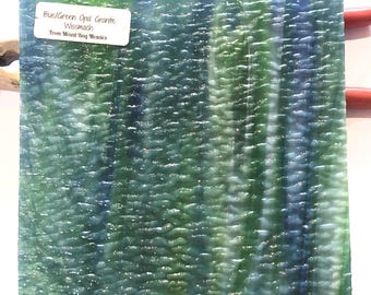 May include: A square piece of blue and green opal granite with a textured surface. The colors blend together, creating a marbled effect. The text on the label reads "Blue/Green Opal Granite Wissmach From Mixed Bag Mosaica."