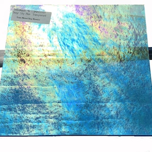 May include: A sheet of iridescent glass with a deep sky blue transparent color. The glass has a textured surface with a variety of colors, including blue, green, yellow, and purple. The text on the label reads "Wismach #6 Luminescent Deep Sky Blue Transparent from Mixed Bag Mosaic".