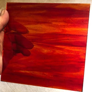 May include: A square piece of stained glass with a fiery red and orange color gradient. The glass has a textured appearance with streaks and variations in color, resembling a sunset or flames. The glass is held by a hand.