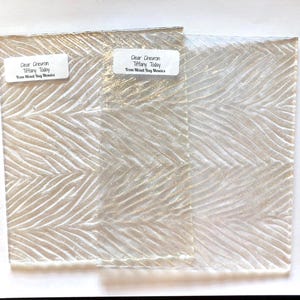 May include: Two clear glass tiles with a chevron pattern and the text "Clear Chevron Tiffany Today From Mixed Bag Mosaics". The tiles have a textured surface and a light, neutral color.
