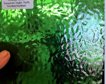 May include: A sheet of green, transparent English Muffle glass with a textured surface. The glass is labeled "Wismach Green Transparent English Muffle from Mixed Bag Mosaics".