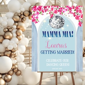 May include: A blue and white wedding sign with pink flowers and a disco ball. The sign reads "Mamma Mia! Lauras Getting Married! Celebrate our Dancing Queen! 14th May 20XX".