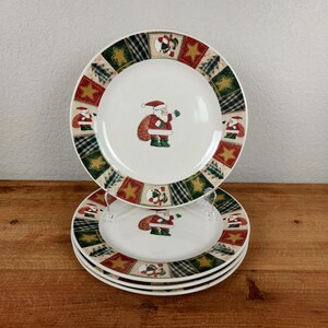 May include: A stack of four white ceramic Christmas plates. Each plate features a central image of Santa Claus and a patterned border with stars, trees, and plaid squares. The plates are on a wooden surface.