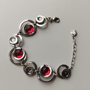 Peut inclure: Un bracelet en métal argenté composé d'une série de maillons circulaires interconnectés à la texture martelée. Le design intègre plusieurs pierres cabochons rondes et translucides de couleur rouge, avec un fermoir mousqueton.
