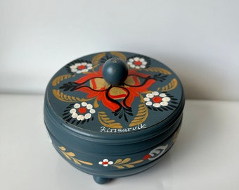 May include: A small, round, blue wooden box with a floral design painted on the lid. The box has three legs and says "Rørosvik" on the side.