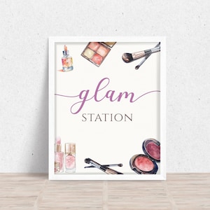 May include: White framed sign with the text "glam STATION" in purple script. The sign is decorated with watercolor illustrations of makeup products, including eyeshadow palettes, lipstick, blush, and makeup brushes.