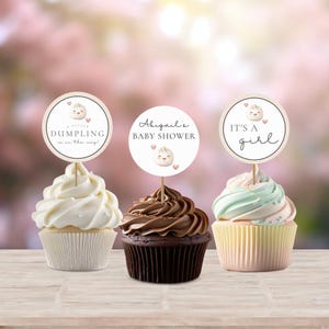 May include: Three cupcakes with different frosting colors and designs. Each cupcake has a round paper topper with a cartoon dumpling face and text. The toppers say "A little dumpling is on the way!" "Abigail's Baby Shower" and "It's a girl!"