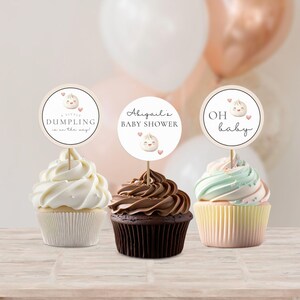 May include: Three cupcakes with white, brown, and green frosting. Each cupcake has a round, white paper topper with a cartoon dumpling and text. The toppers say "A little dumpling is on the way!" "Abigail's Baby Shower" and "Oh baby".