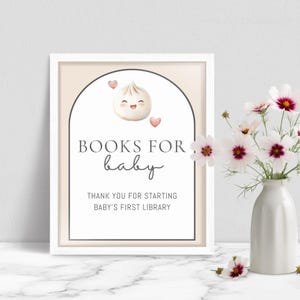 May include: A framed sign with the text "BOOKS FOR baby" and a cartoon dumpling illustration. The sign also includes the text "THANK YOU FOR STARTING BABY'S FIRST LIBRARY". A vase of white and pink flowers is to the right.