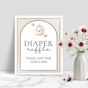 May include: A framed sign with the words "DIAPER raffle" and "PLEASE LEAVE YOUR TICKETS HERE". The sign features a cartoon dumpling with hearts. A vase of white and pink flowers sits to the right.