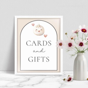 May include: A framed sign with the words "CARDS and GIFTS" in a whimsical font. The sign features a cartoon dumpling with hearts. A vase of white and pink flowers sits to the right of the sign.