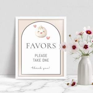May include: A framed sign with the word "FAVORS" in a serif font, above the text "PLEASE TAKE ONE" and "thank you!". The sign features a cartoon dumpling with a smiling face and two pink hearts. A vase of flowers is to the right.